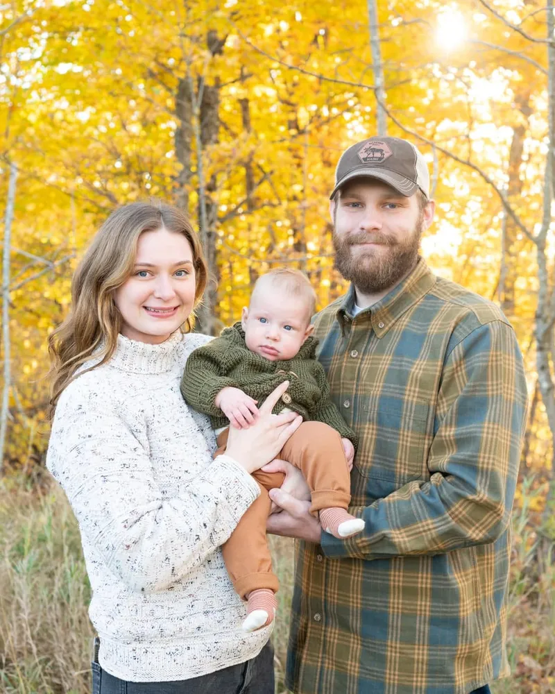 A mother and father holding their baby in front of golden autumn birch trees backlit by the sun.