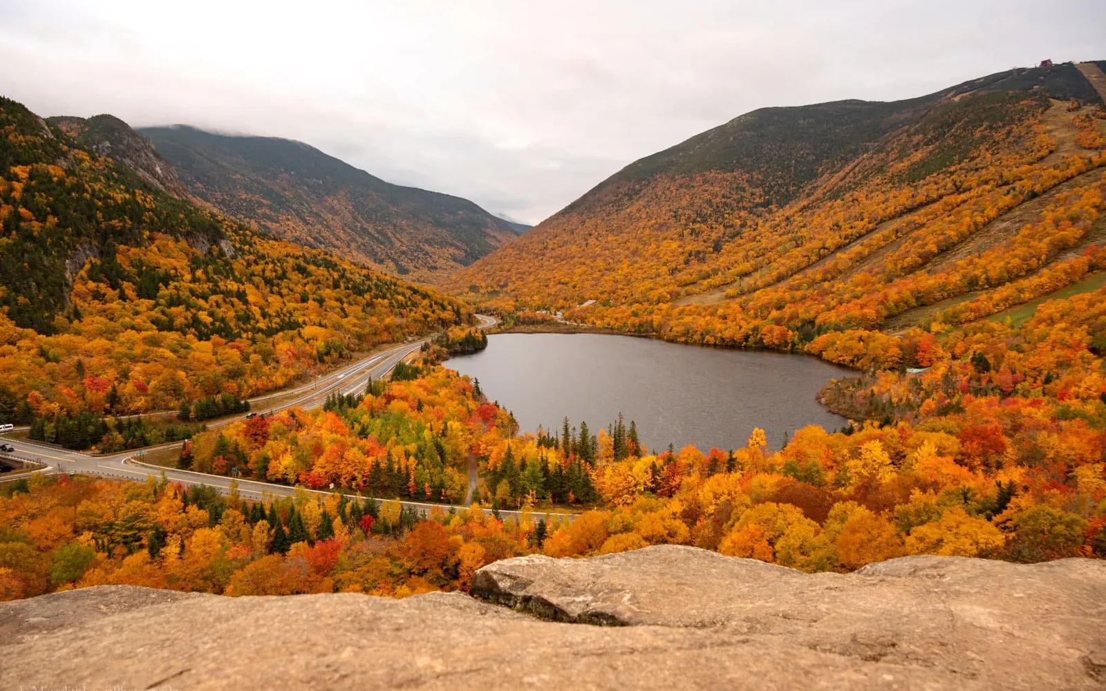 An elevated view of a New Hampshire notch in the White Mountains, with peak autumn foliage covering the mountainsides above a lake and a winding road.