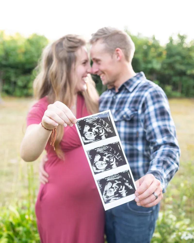 An expectant couple standing in a sunlit field, holding up a strip of black-and-white ultrasound photos.